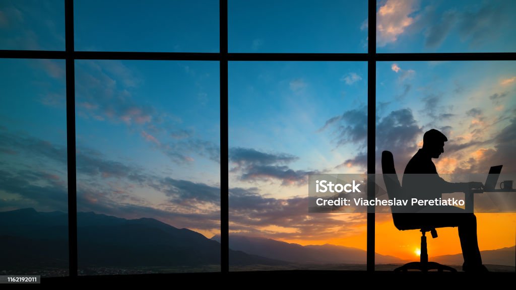 The man working at the table near a window on a mountain sunset background Window Stock Photo