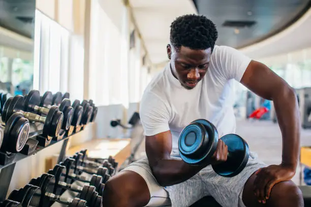 Photo of Young African American man sitting and lifting a dumbbell with the rack at gym