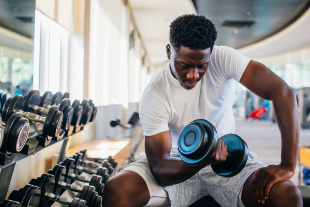 年輕的非洲裔美國男子坐在健身房的架子上,舉起一個啞鈴 - 舉重訓練 個照片及圖片檔