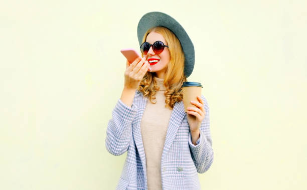 istock Portrait happy smiling young woman with smartphone using voice command recorder or calling, wearing coat jacket, round hat on wall background 1162201729