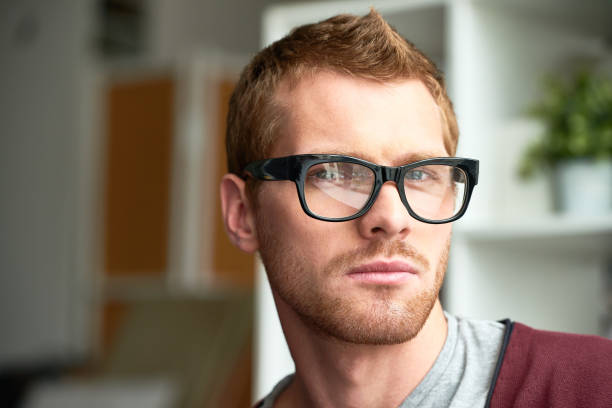 Headshot of young bearded man in black glasses looking seriously at camera