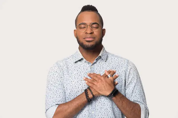 Mixed race man in glasses isolated on grey white studio background, guy closed eyes feels grateful for love and support holds hands on chest show sincere feelings come from heart, concept of gratitude