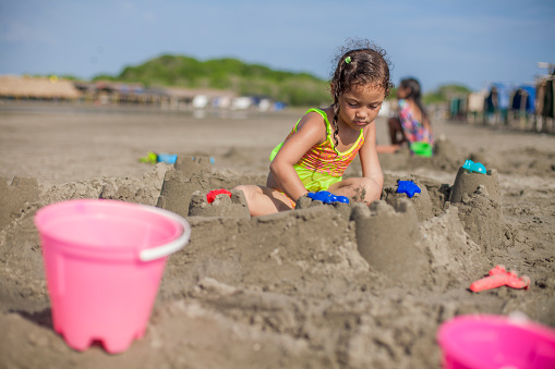 Little girl of Latin race, sitting on the beach, making sandcastles, while having a sunny day, smiling and having fun