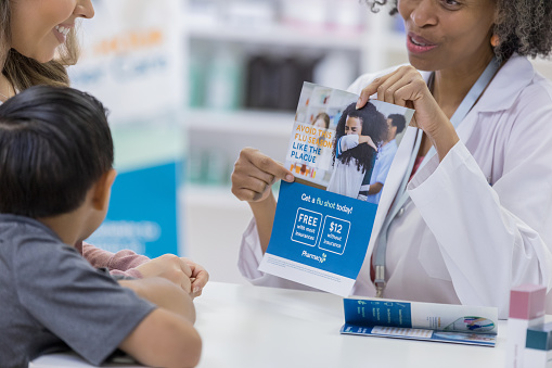 Female pharmacist discusses flu prevention with a customer and the customer's son. The pharmacist is showing them a flu vaccine informational flyer.