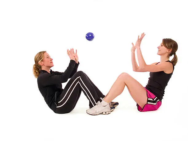Full length image of two adult women exercising together by throwing a small ball/weight back and forth while holding themselves at the top of a sit-up position; copy space 