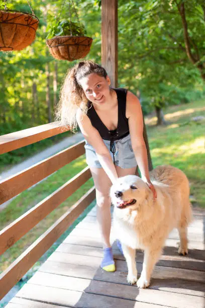 Young smiling woman petting one happy white great pyrenees dogs outside at home porch of log cabin