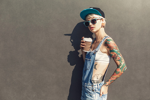 Young alternative girl wearing cap and sunglasses standing isolated on grey wall on the city street holding cup drinking hot coffee looking camera pensive