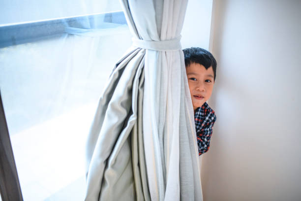 Young Taiwanese boy playing hide and seek with the help of a curtain in the interior of his contemporary home.