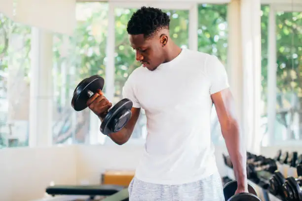 Photo of Young African American man standing and lifting a dumbbell with the rack at gym