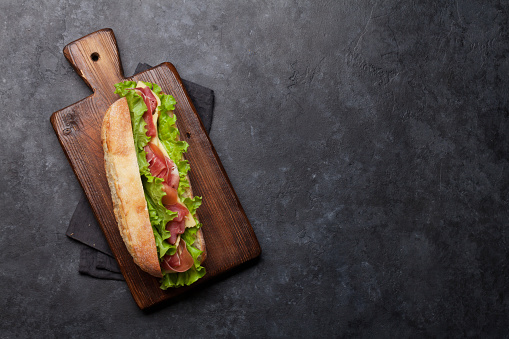 Fresh submarine sandwich with prosciutto ham, cheese and lettuce on dark stone background. Top view with copy space for your text
