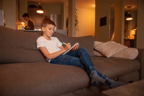 istock Two people, one boy, writing in notebook, while sitting in couch. Man behind in kitchen, out of focus. 1162278370