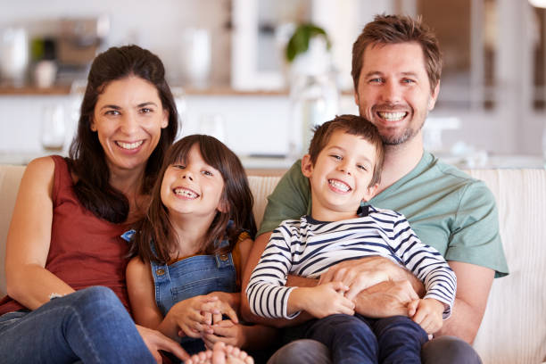 pareja blanca de adultomedio y sus dos hijos pequeños sentados en un sofá en casa sonriendo a la cámara - family white family with two children cheerful fotografías e imágenes de stock