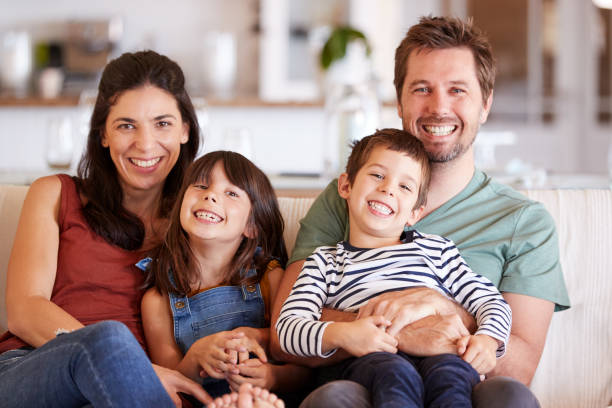 coppia bianca adulta media e i loro due bambini piccoli seduti su un divano a casa sorridenti alla telecamera - four people foto e immagini stock