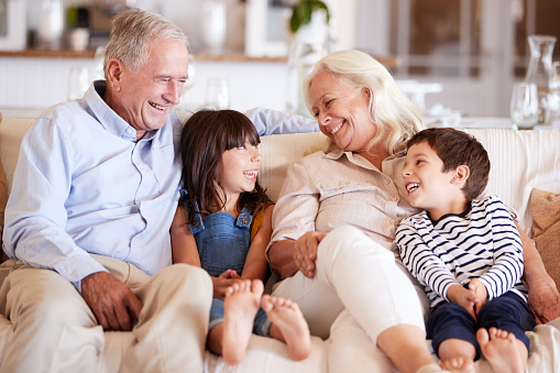 Pareja blanca de la tercera edad y sus nietos sentados en un sofá juntos sonriendo el uno al otro photo