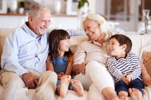 white senior couple and their grandchildren sitting on a sofa together smiling at each other - grandparent grandfather grandmother child imagens e fotografias de stock