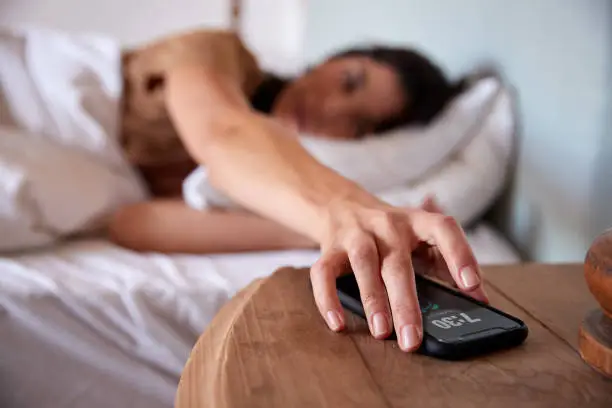 Mid adult woman lying in bed, reaching out to smartphone on the bedside table in the foreground