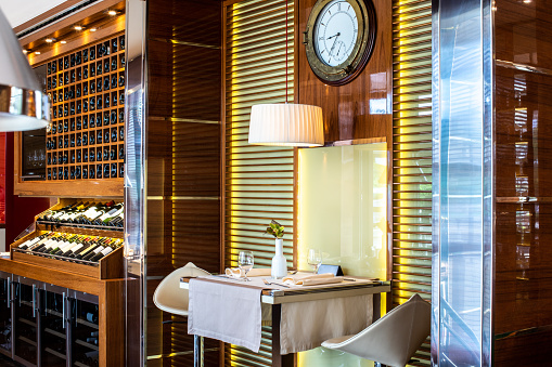 A two seated table set next to a wine display in a high profile restaurant. A nautical clock and a lamp are hanging above.