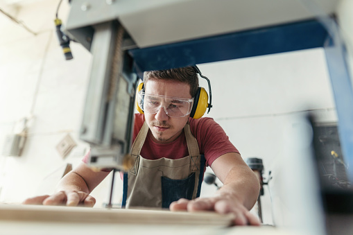 Young crafts worker in protective workwear cutting wooden piece with grinding machine