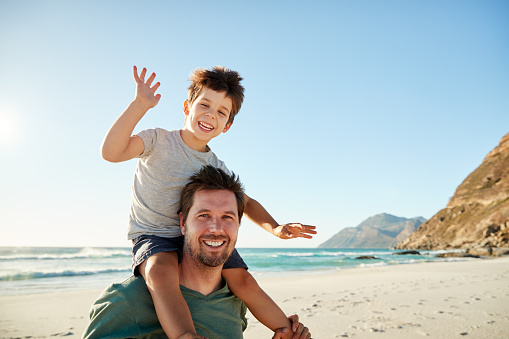 Mid adult white man on a beach, his four year old son sitting on his shoulders waving, close up
