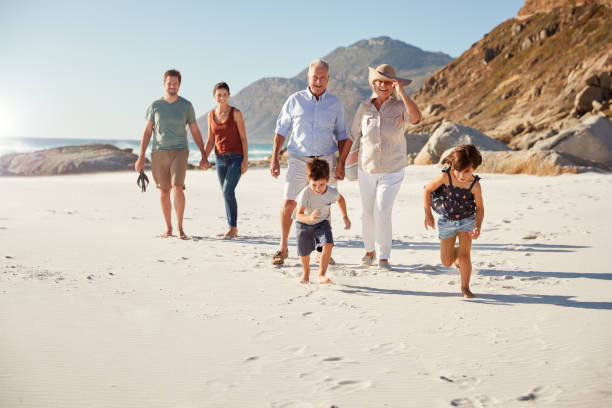 日当たりの良いビーチで一緒に歩く3世代の白人家族、前方を走る子供たち - 旅行 写真 ストックフォトと画像