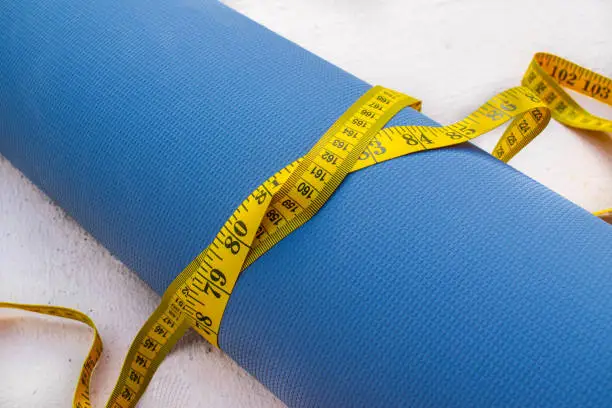 Photo of yoga weightloss symbolized by a measuring yellow tape tied around a blue yoga mat on a white background