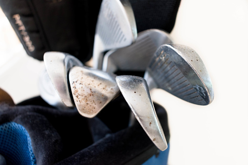 Close up of rusty golf clubs, irons, wedges and a driver with a black cover in a blue and black back with an off white background.