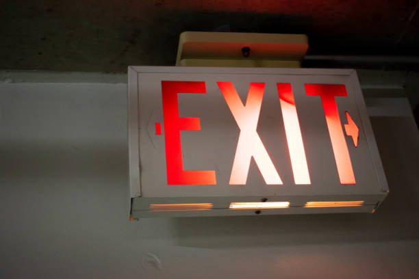 istock A red, orange, white glowing exit sign with an arrow pointing to the right at night in a hallway. 1162303695