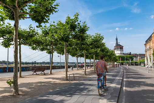 Dusseldorf, Germany - June 25 2019: Man cycles by a row of trees along the River Rhine in Dusseldorf.