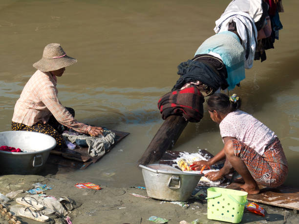 Amarapura, Mandalay, Myanmar ( Burma) - January 15th 2016 : Two women crouching washing their clothes at the shores of the Taungthaman lake. The laundry is in a zinc bucket, brushed on boards and put it on a wooden bar.