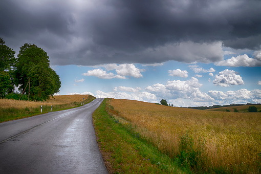 Dark grey threatening storm clouds gathering over a country road running through open fields to disappear over the hill towards blue sunny sky with white clouds in the distance