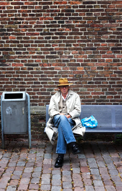 Groningen, Netherlands: A senior man with a fedora and raincoat relaxing on a bench against a brick wall in downtown Groningen.