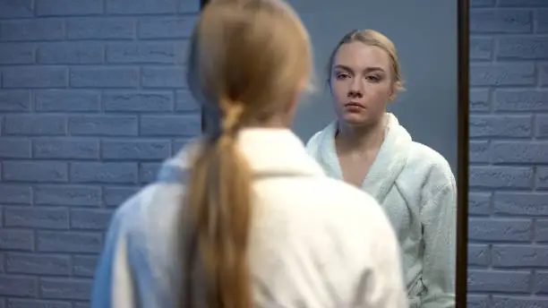 Unhappy young lady in bathrobe suffering stress looking in mirror, soul pain