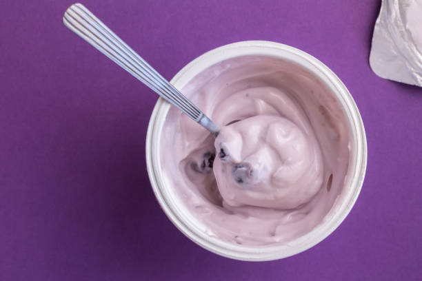 tazza di yogurt con yogurt alle bacche blu, cucchiaio e coperchio in foil isolati su sfondo viola - yogurt container foto e immagini stock