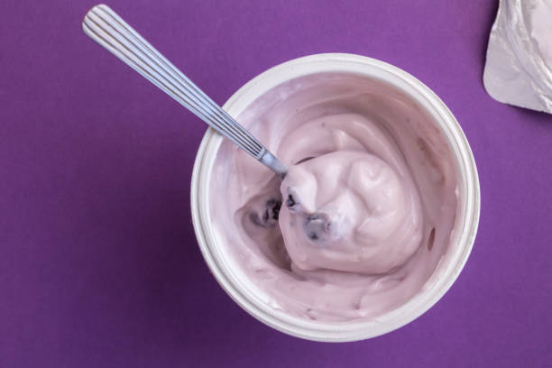 Yogurt cup with blue berry yoghurt, spoon and foil lid isolated on purple background Yogurt cup with blue berry yoghurt, spoon and foil lid isolated on purple background - top view photograph yoghurt stock pictures, royalty-free photos & images