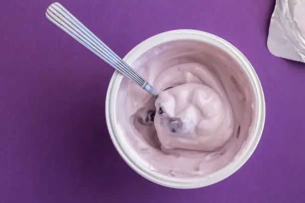Photo of Yogurt cup with blue berry yoghurt, spoon and foil lid isolated on purple background