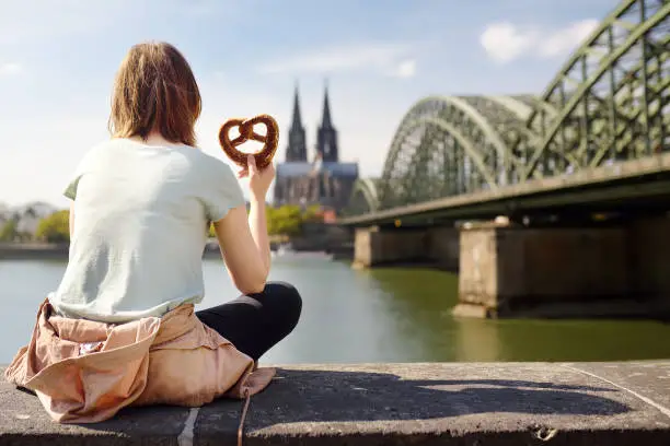 Young woman eats traditional pretzel sitting on embankment of Rhine on background of Cologne Cathedral and Hohenzollern Bridge in Koel, Germany. Tourism and travel by Germany.