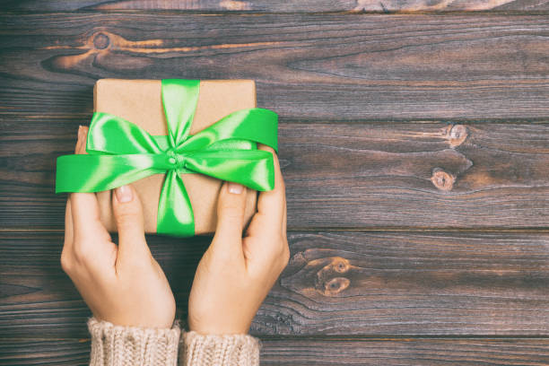 les mains de femme donnent la valentine enveloppée ou le cadeau fait à la main de vacances dans le papier avec le ruban vert. boîte de présentation, décoration du cadeau sur la table en bois blanche, vue supérieure avec l'espace de copie - isolated on white bow gift homemade photos et images de collection