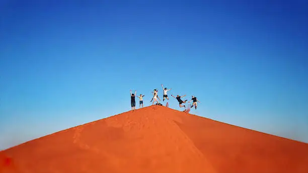 Group of 7 people with kids, teenagers and adults jumping on a triangular shaped sand dune Red Sand dune fun NamibRand Namibia Africa