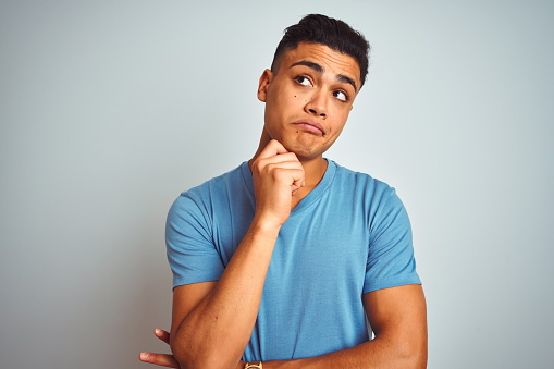 Young brazilian man wearing blue t-shirt standing over isolated white background with hand on chin thinking about question, pensive expression. Smiling with thoughtful face. Doubt concept.