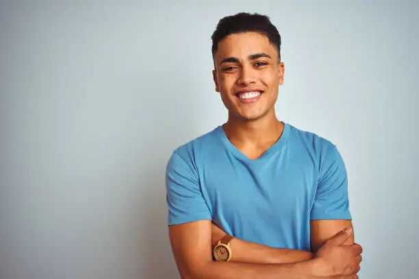 Photo of Young brazilian man wearing blue t-shirt standing over isolated white background happy face smiling with crossed arms looking at the camera. Positive person.