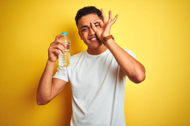 giovane brasiliano che tiene in mano una bottiglia d'acqua in piedi su uno sfondo giallo isolato con il viso felice sorridente facendo un segno ok con la mano sull'occhio che guarda tra le dita - drinking men water bottle foto e immagini stock