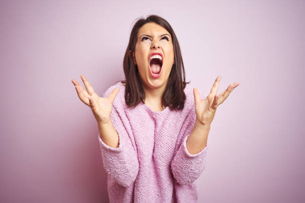 ピンクの孤立した背景の上にセーターを着ている若い美しいブルネットの女性は狂気と怒鳴り、攻撃的な表情と腕を上げて叫ぶ。欲求不満の概念。 - frustration ストックフォトと画像