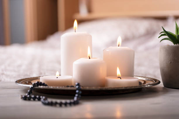 Composition of white burning candles on bedside table in light cozy bedroom interior. Selective focus. With space for text.