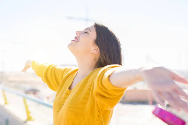 Beautiful young woman smiling with closed eyes and open arms enjoying the sunlight on a sunny day. Happy sunbathe