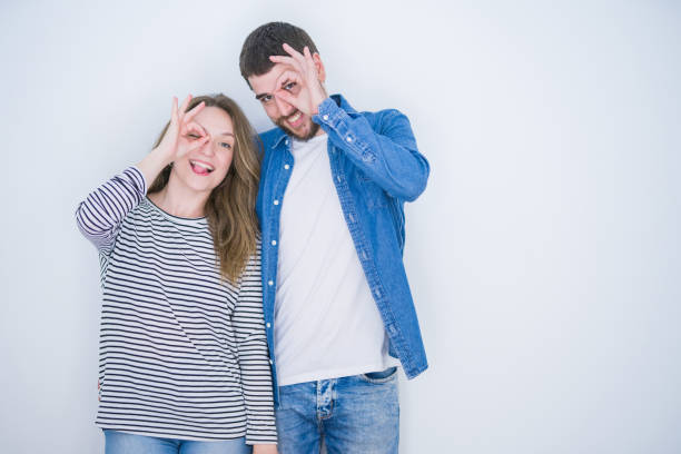 istock Young beautiful couple standing together over white isolated background doing ok gesture with hand smiling, eye looking through fingers with happy face. 1162360514