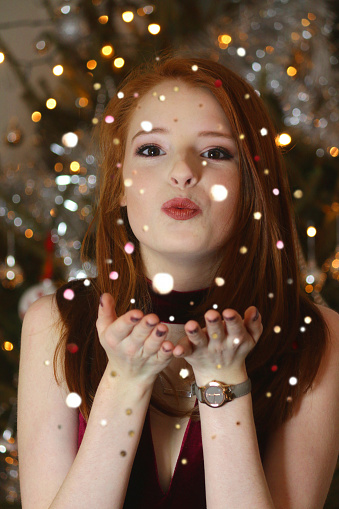 Stock photo of beautiful young woman with red hair / pretty teenage girl 14 / 15 / 16 years old with ginger hair at Christmas New Year's Eve party, blowing sparkling confetti glitter, wearing red velvet party dress, Christmas tree fairy lights background