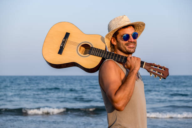 A young woman in a cowboy hat plays the guitar on the shore of the blue sea on a sunny day. The beach is quiet and peaceful. The guitarist is giving a concert by the ocean. Guitar lessons.