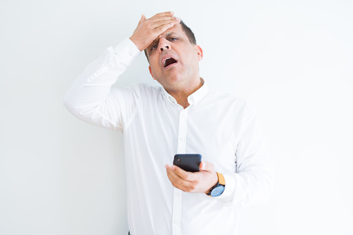 Middle age man using smartphone over white wall stressed with hand on head, shocked with shame and surprise face, angry and frustrated. Fear and upset for mistake.