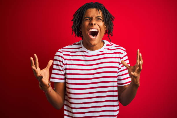 istock Young afro man with dreadlocks wearing striped t-shirt standing over isolated red background crazy and mad shouting and yelling with aggressive expression and arms raised. Frustration concept. 1162383699