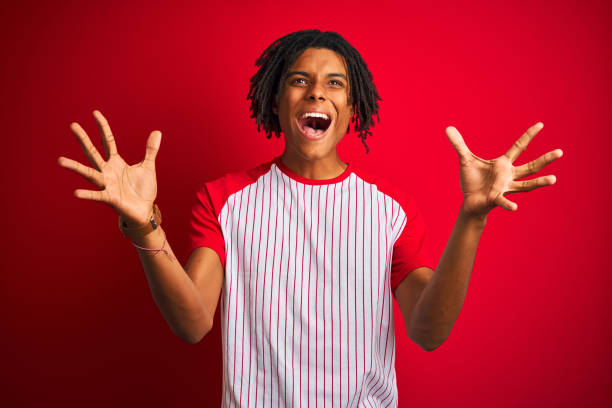 istock Afro man with dreadlocks wearing striped t-shirt standing over isolated red background celebrating crazy and amazed for success with arms raised and open eyes screaming excited. Winner concept 1162383811